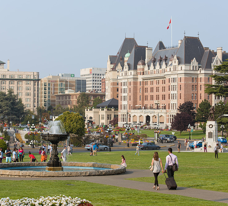 People enjoy sightseeing tours, strolling across a grassy park with a fountain, in front of a large historic hotel topped with a Canadian flag and surrounded by city buildings.