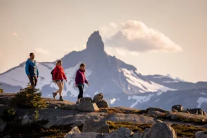 Three people in hiking clothes walk on a rocky trail with a snow-capped mountain and cloudy sky in the background.