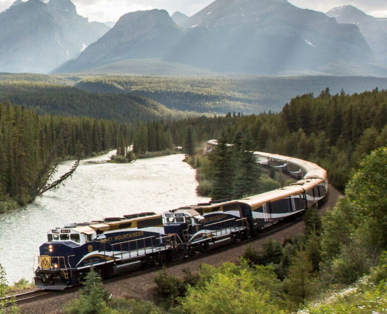 A passenger train labeled "Rocky Mountaineer" embarks on a scenic tour along a riverside track through a forested mountain landscape under a cloudy sky, perfect for unforgettable sightseeing tours.