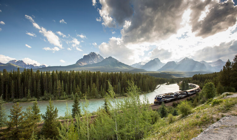 A train travels along tracks beside a river, surrounded by dense forest and mountain peaks under a partly cloudy sky—the perfect setting for unforgettable sightseeing tours.
