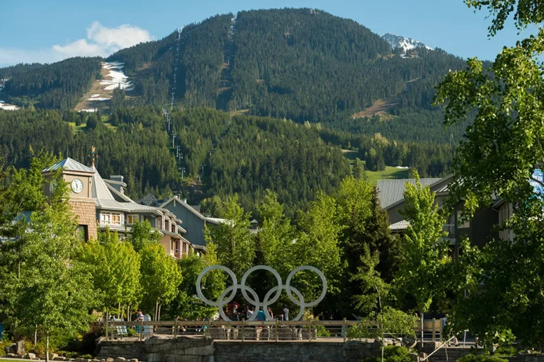 A mountain with ski runs in the background, buildings in the foreground, and large Olympic rings sculpture in a park area surrounded by trees—an ideal spot for sightseeing tours