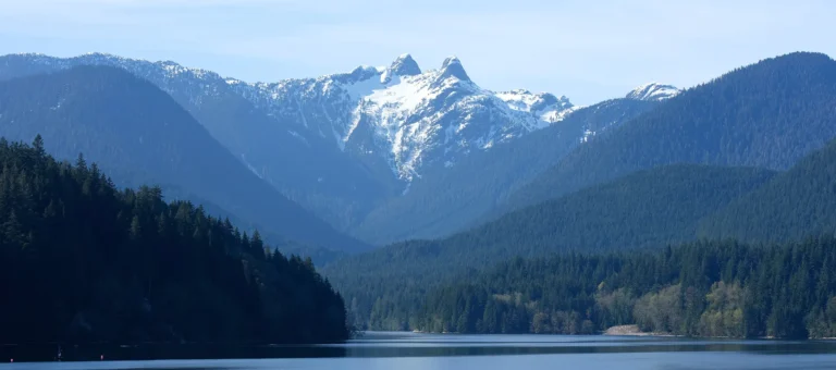 Snow-capped mountain peaks rise above forested hills and a calm lake under a clear sky, making it the perfect setting for unforgettable sightseeing tours.