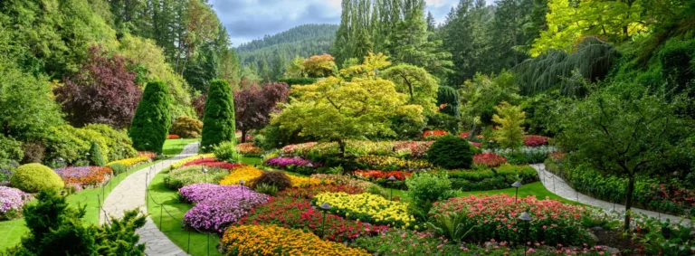 A landscaped garden with winding stone paths, colorful flower beds, green shrubs, and tall trees, set against a backdrop of forested hills under a partly cloudy sky.