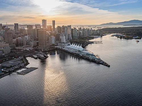 Aerial view of downtown Vancouver at sunset with the Canada Place white sail roof and waterfront, surrounded by water and distant mountains.