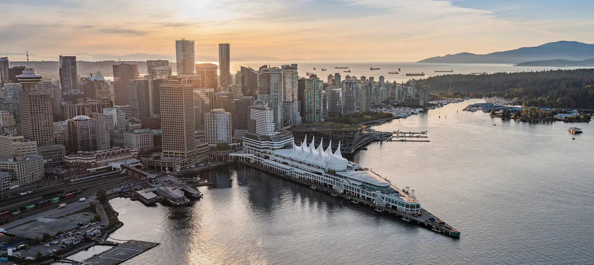 Aerial view of downtown Vancouver at sunset, featuring Canada Place with its white sails—an iconic stop on many sightseeing tours—surrounded by high-rise buildings and water with ships anchored offshore