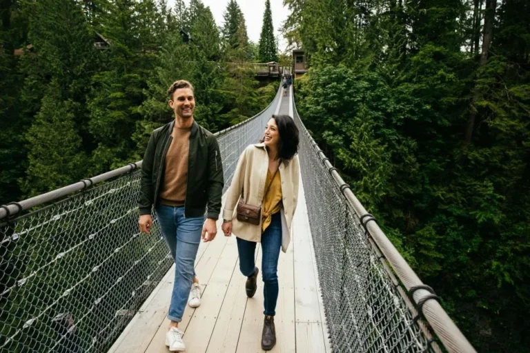 Two people walk and smile on a suspension bridge surrounded by dense green forest.