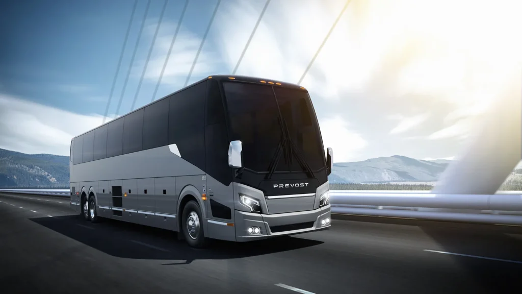A large, modern Prevost coach bus drives on a bridge with mountains and a bright sky in the background.