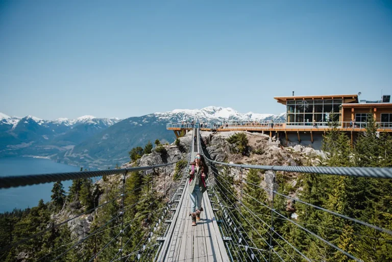 A person enjoys a sightseeing tour, walking on a suspension bridge surrounded by mountains and trees, with a modern building visible on the right under a clear blue sky