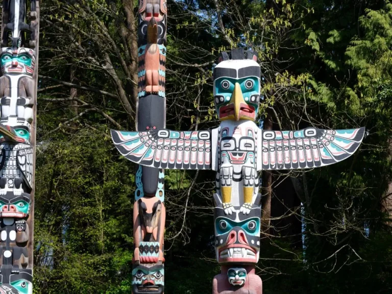 A brightly painted Indigenous totem pole featuring a large bird with outstretched wings stands among trees, making it a striking highlight for sightseeing tours in this scenic outdoor setting