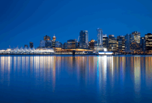 City skyline at night with illuminated buildings reflecting on calm water under a clear, dark blue sky.