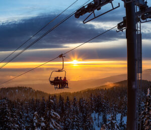 A ski lift with several people rises over a snowy forested mountain landscape at sunset, with the sun low on the horizon and clouds in the sky.