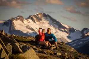 Two people sit on rocky ground taking a selfie with snow-capped mountains in the background under a partly cloudy sky.