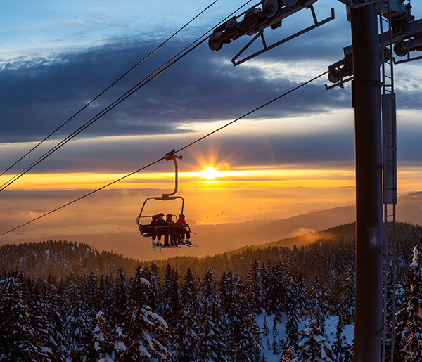 A ski lift with several people rises over a snowy forested mountain landscape at sunset, with the sun low on the horizon and clouds in the sky.