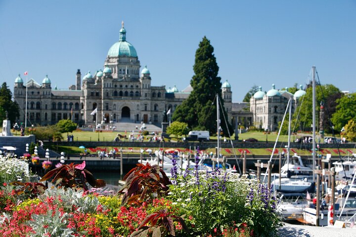 The image shows the British Columbia Parliament Buildings behind a harbor with boats, colorful flowers in the foreground, and people on the lawn under a clear blue sky