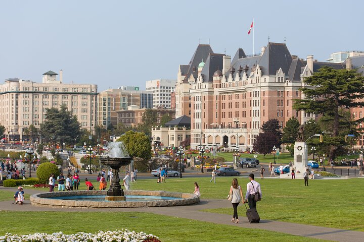 People walk and gather near a fountain in a park, with historic buildings and a large hotel in the background under a clear sky