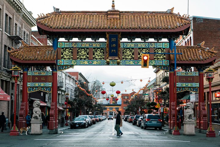 A large, ornate Chinese gate spans a busy city street with cars parked along both sides and pedestrians crossing underneath. Red lanterns hang above the street