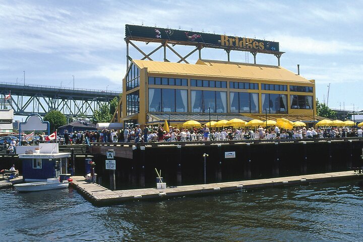 Waterfront restaurant with yellow accents and outdoor seating under yellow umbrellas, crowded with people, with a bridge and boats visible in the background