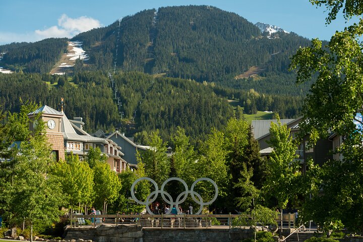 The Olympic rings monument stands in a landscaped area with buildings and forested mountains in the background on a sunny day, making it a popular stop for Sightseeing Tours Whistler