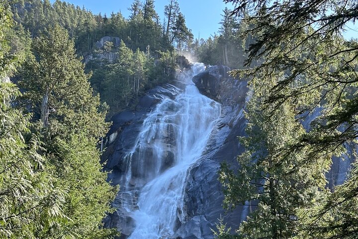 Water cascades down a rocky cliff surrounded by dense green trees under a clear blue sky—a perfect spot for Sightseeing Tours Whistler
