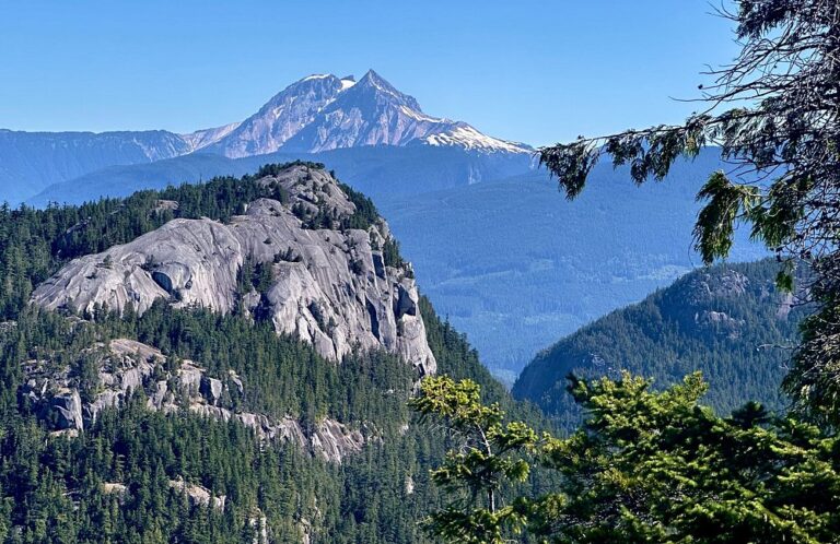 A rocky mountain peak rises above a forested landscape, with a snow-capped mountain in the background under a clear blue sky—the perfect scene for unforgettable Sightseeing Tours Whistler