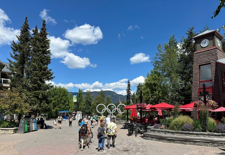 People walk through a plaza with the Olympic rings sculpture, surrounded by trees, outdoor seating with red umbrellas, and a clock tower under a sunny blue sky—a perfect spot for Sightseeing Tours Whistler