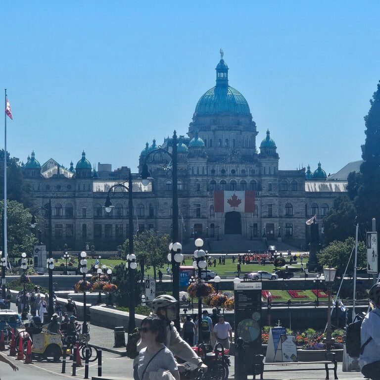 The British Columbia Parliament Buildings in Victoria, Canada, with a large Canadian flag displayed on the front and people gathered in the foreground on a sunny day