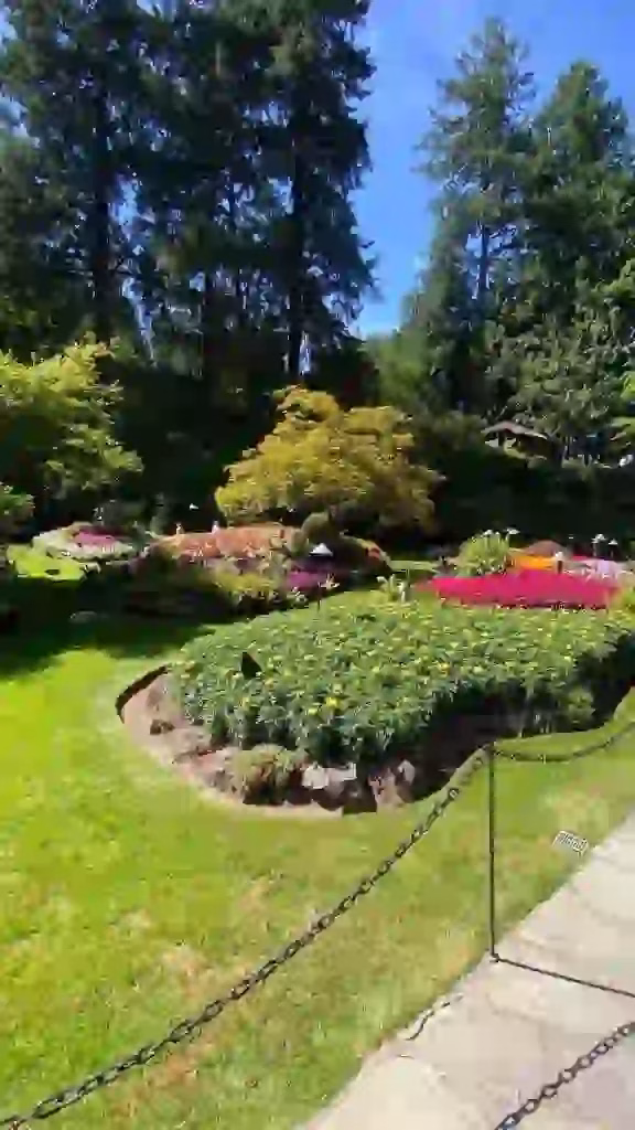 A landscaped garden with flower beds, green shrubs, and tall trees in the background under a clear blue sky. A chain barrier borders the sidewalk in the foreground, reminiscent of scenes from a Butchart Gardens Tour.