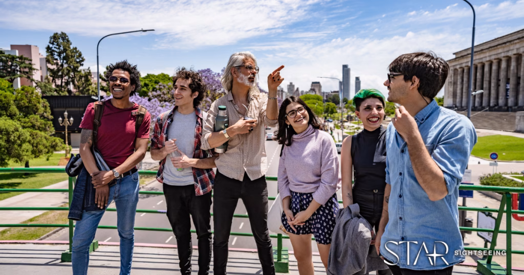 A group of six people stand on a city bridge, smiling and talking, with trees, buildings, and a road in the background.