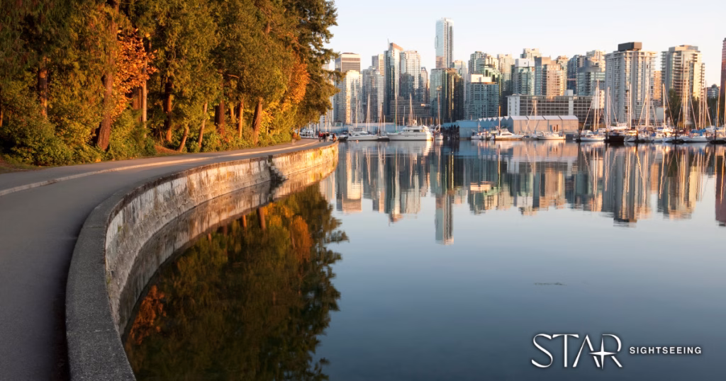 A paved seawall path curves beside calm water, reflecting moored boats and high-rise buildings, with trees on the left and city skyline in the background.