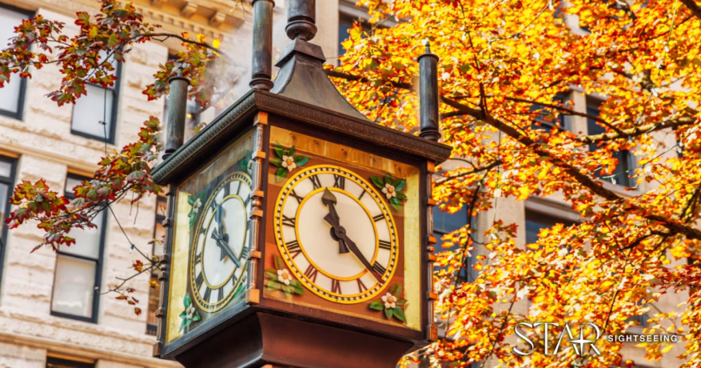 A close-up of a historic steam clock surrounded by autumn leaves, set against the background of an older building.