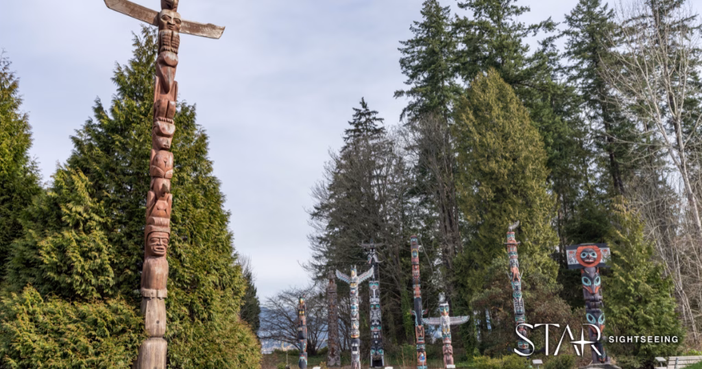 A row of totem poles stands among tall evergreen trees in an outdoor park setting under a cloudy sky.