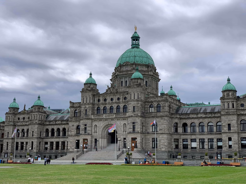 A large stone building with a central dome and green copper roofs stands under a cloudy sky, with flags and people near the entrance—a hallmark stop on any sightseeing tour in Victoria.