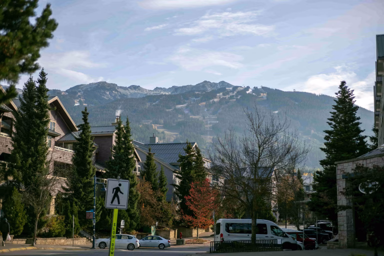 A street scene with houses, trees, parked vehicles, and a pedestrian crossing sign, set against a backdrop of mountains under a partly cloudy sky.