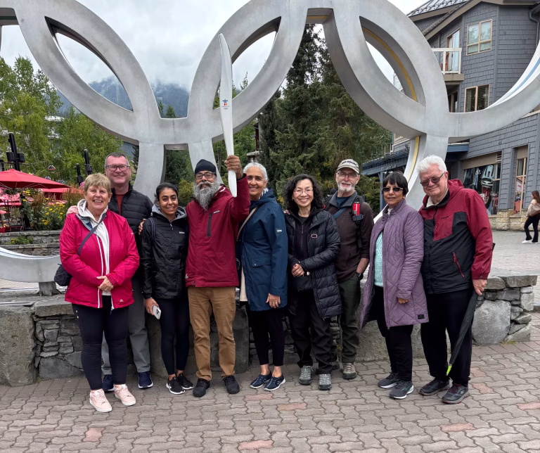 A group of nine adults poses in front of large Olympic rings outdoors, with one person holding up a torch-shaped object. Trees and buildings are visible in the background