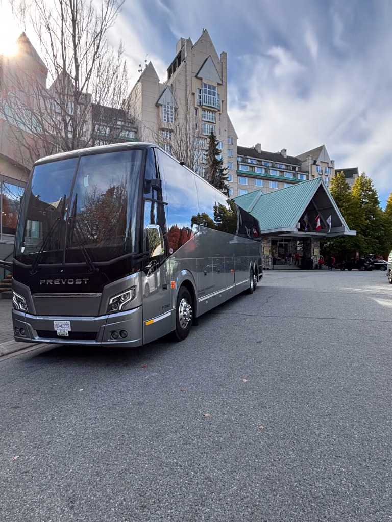 A large grey Prevost tour bus is parked in front of a multi-story hotel with steep roofs, under a partly cloudy sky.