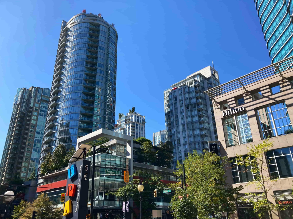 Modern high-rise buildings and a Hilton hotel under a clear blue sky, with trees and colorful parking signs in the foreground—an inviting scene for tours in Downtown Vancouver.