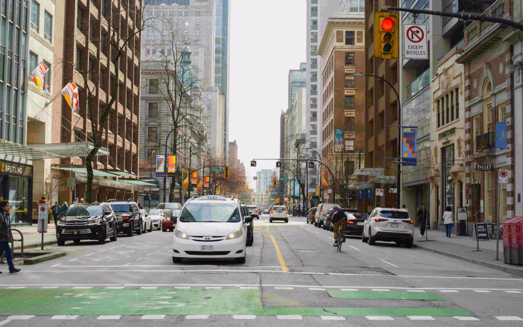City street scene with cars, a taxi, a cyclist, and pedestrians near tall buildings; a traffic light is red and a no parking sign is visible—perfect for capturing the vibrant atmosphere of a Vancouver City Tour.