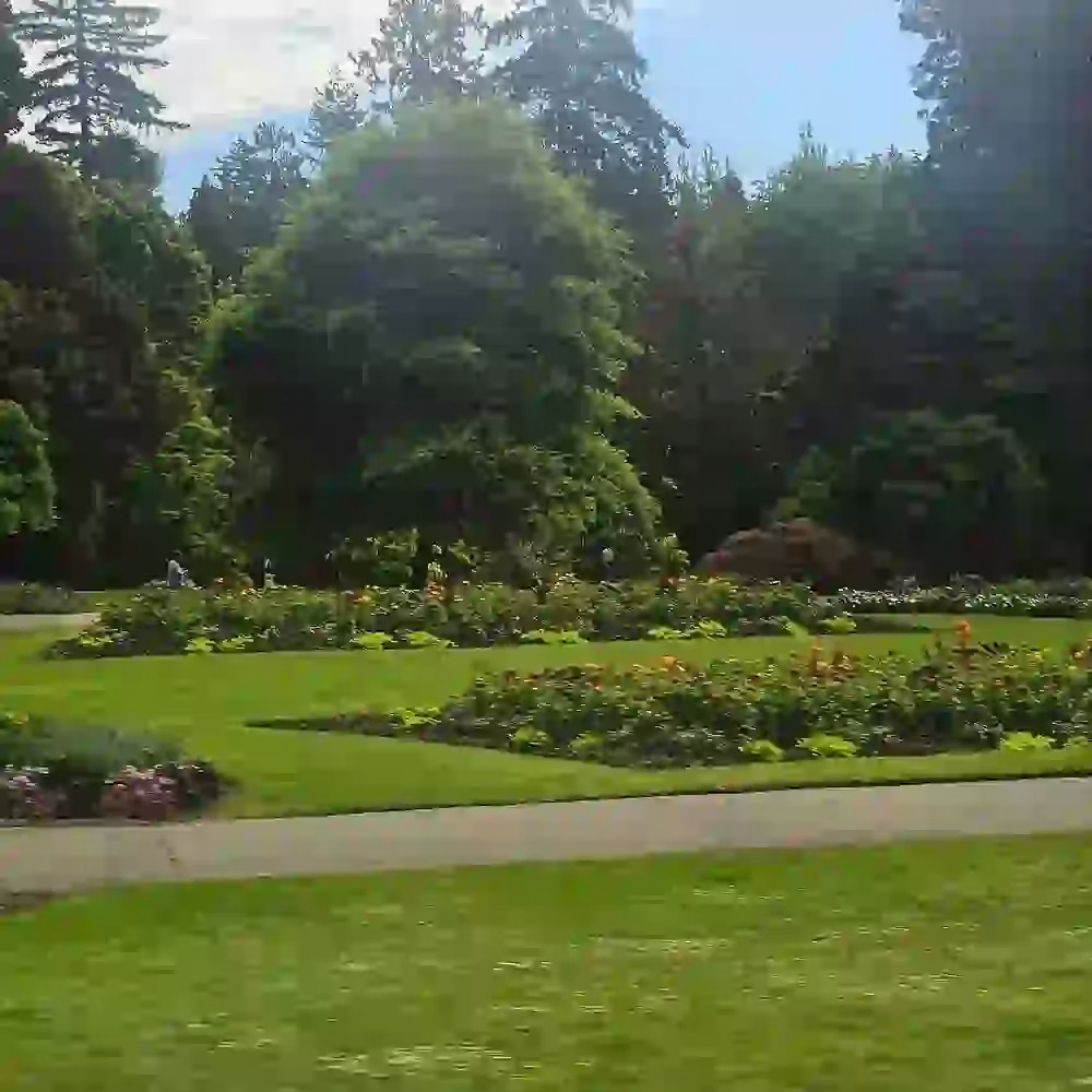 A landscaped garden with green lawns, flower beds, and tall trees in the background under a partly cloudy sky.