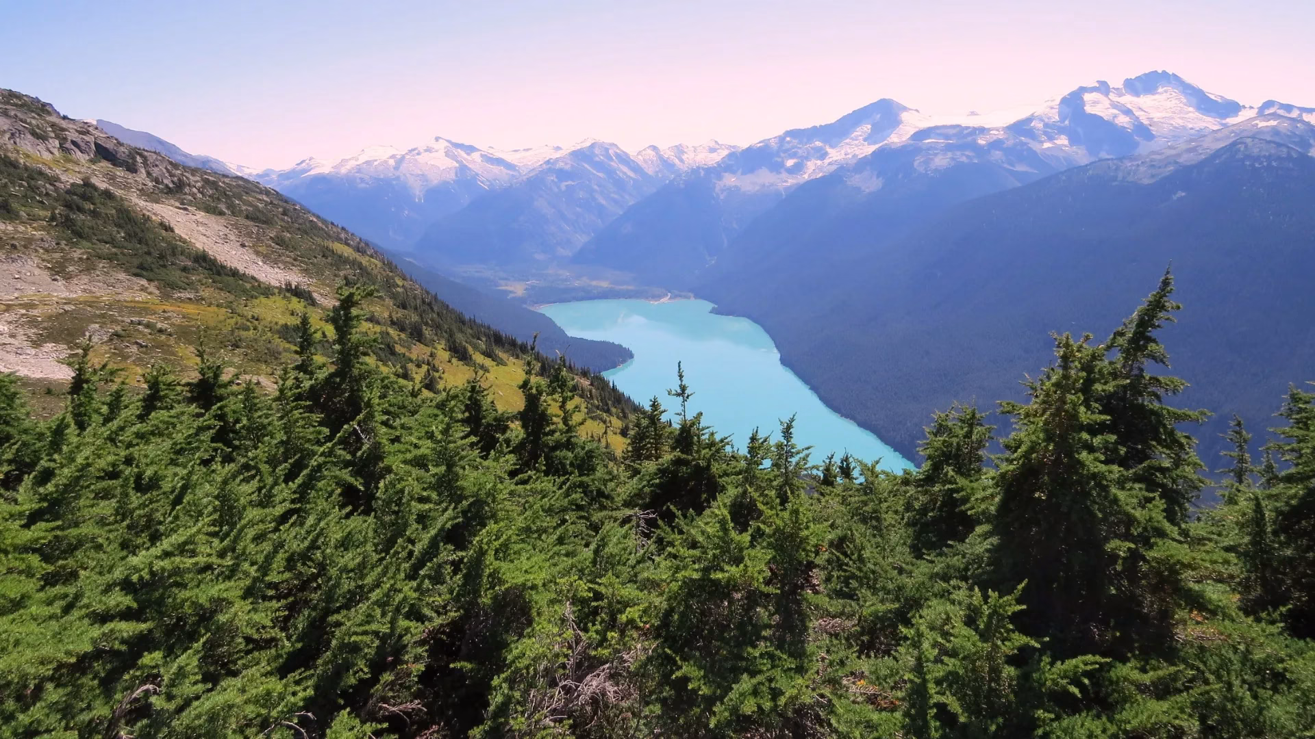 A turquoise lake winds through a valley surrounded by evergreen trees and snow-capped mountains under a clear sky