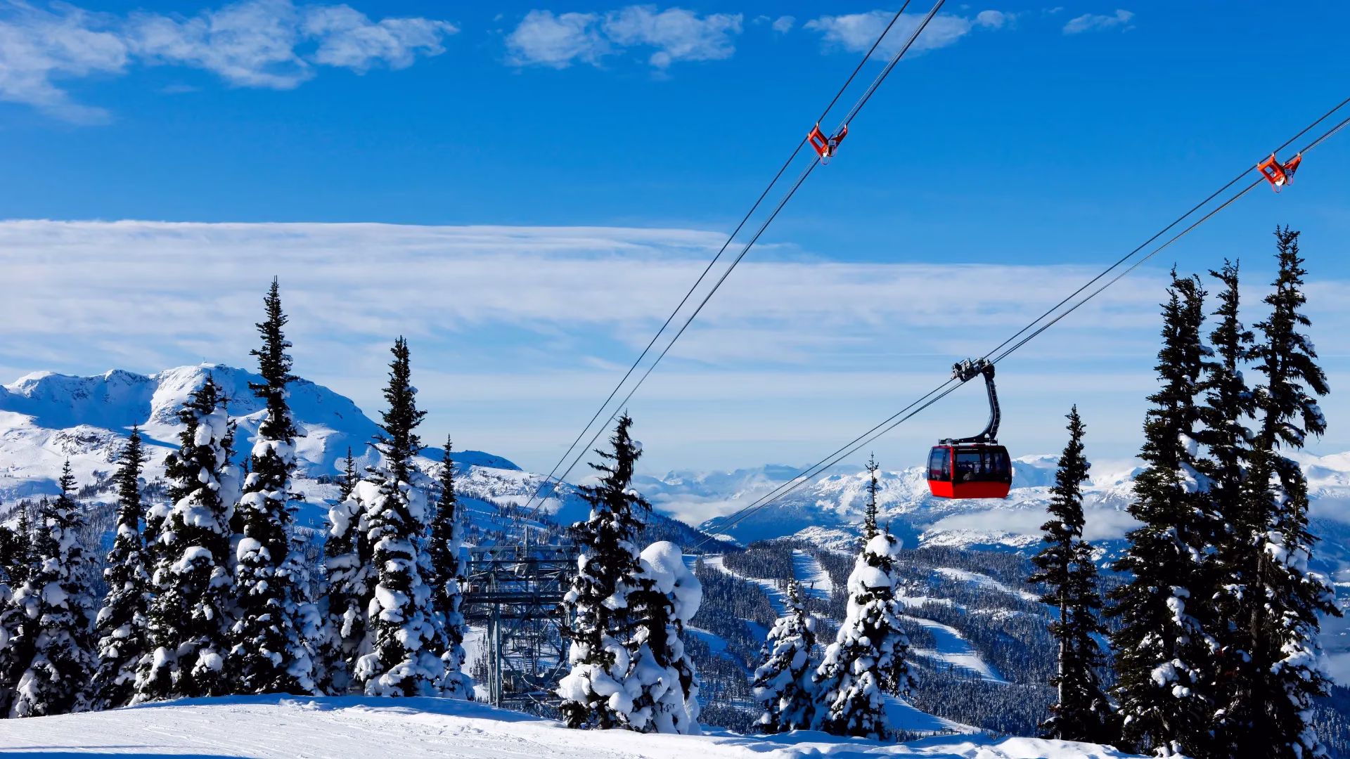 A red gondola travels above snowy pine trees in a mountainous winter landscape under a blue sky