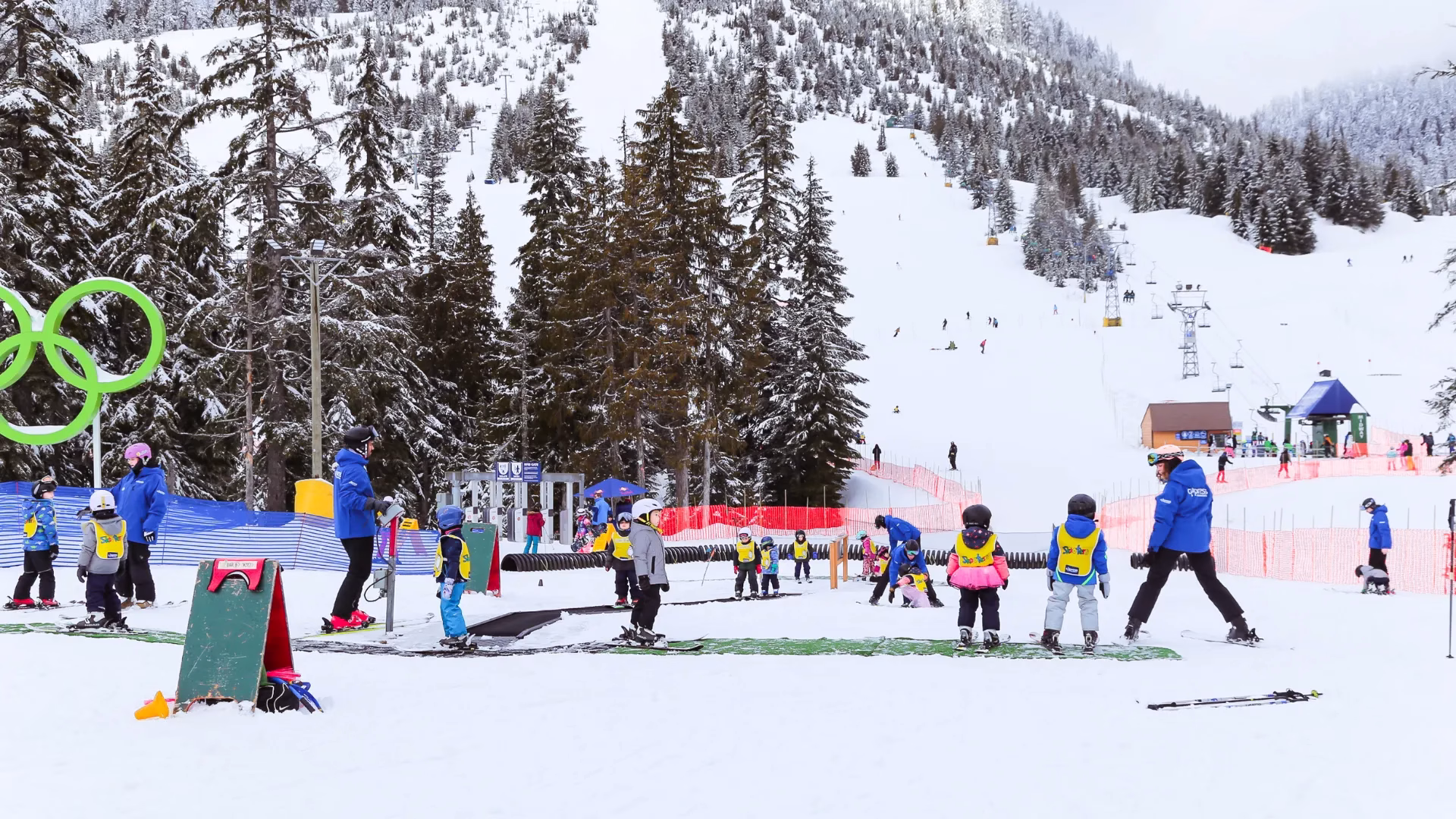 Children and instructors in colorful ski gear gather for a lesson on a snowy slope at a ski resort, with pine trees and a ski lift in the background