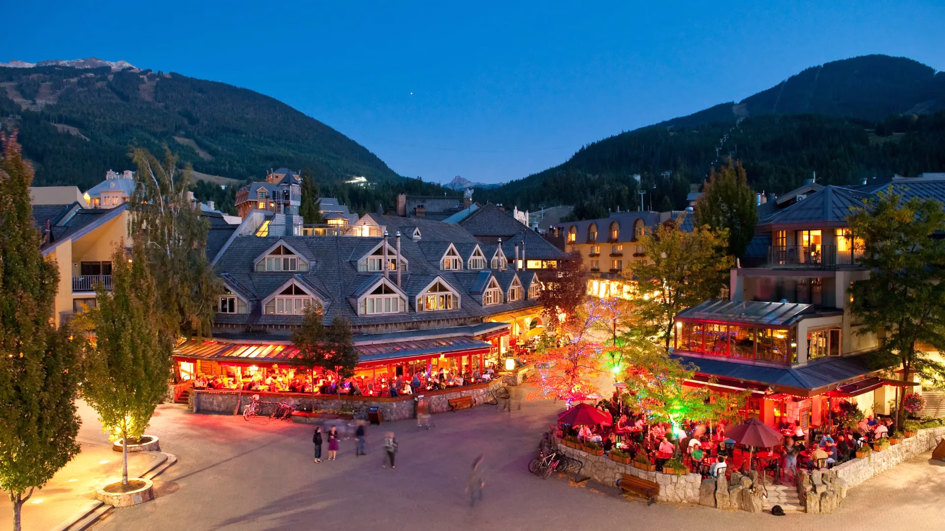 An evening view of a lively village square with lit restaurants, outdoor seating, people walking, and mountains in the background