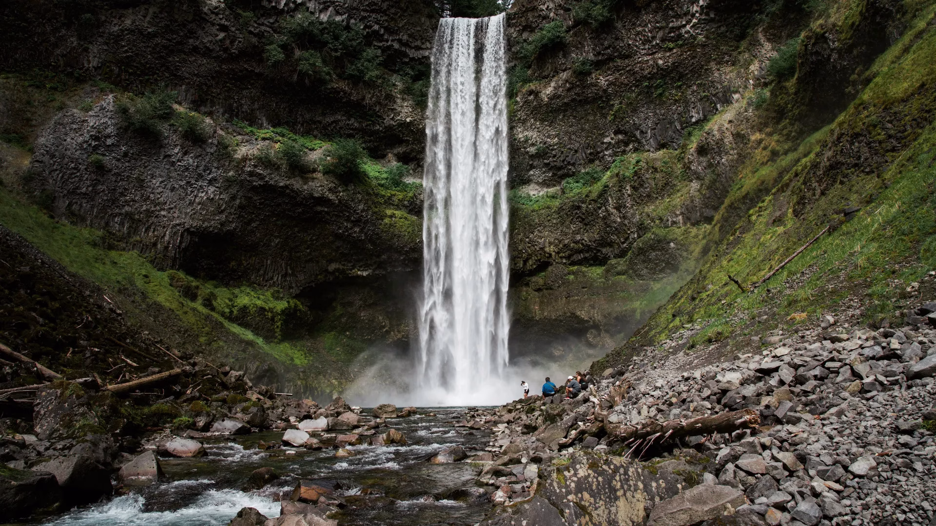 A tall waterfall cascades down a rocky cliff into a pool surrounded by stones and greenery, with a few people standing near the base