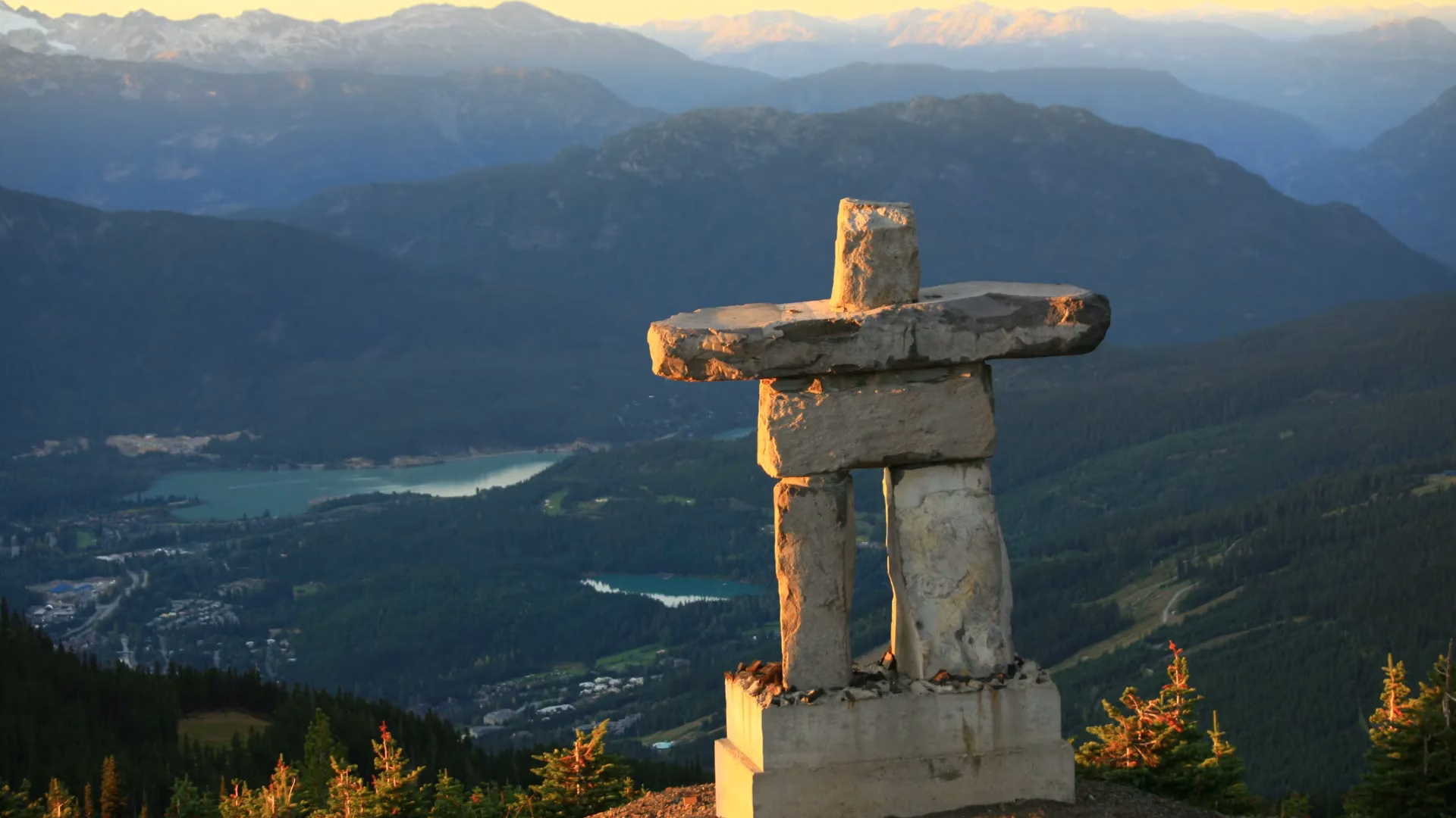 Stone inuksuk statue stands on a mountain overlooking a green valley, forests, lakes, and distant mountain ranges under soft sunlight