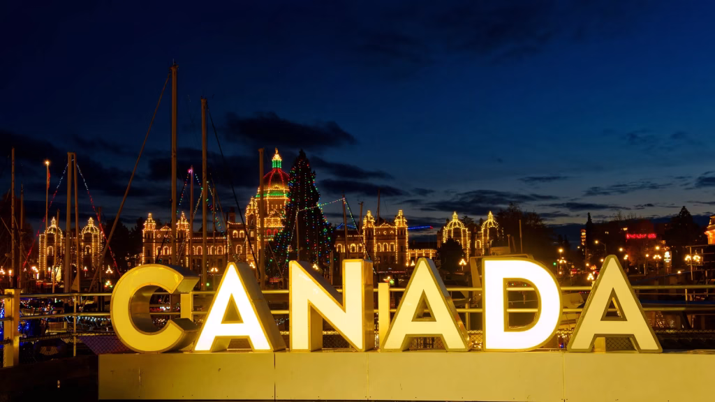 Large illuminated "CANADA" sign in front of buildings decorated with holiday lights at night, with a dark blue sky and silhouetted structures in the background.