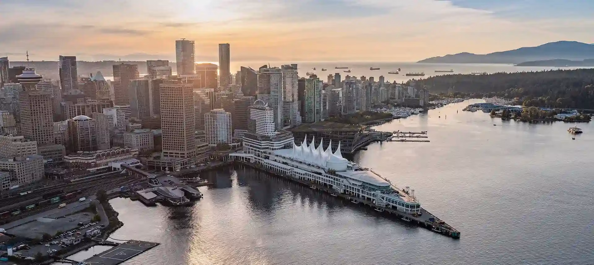 Aerial view of downtown Vancouver with Canada Place, high-rise buildings, and waterfront at sunset. Ships are visible in the harbor