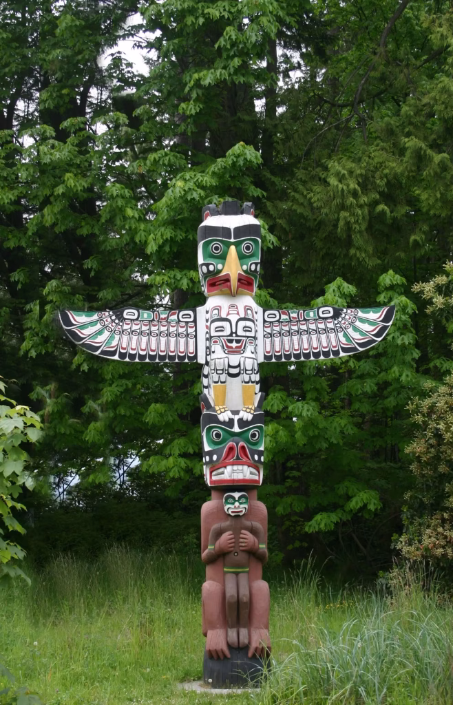 A tall, colorful totem pole with bird and animal carvings stands in front of dense green trees and grass.