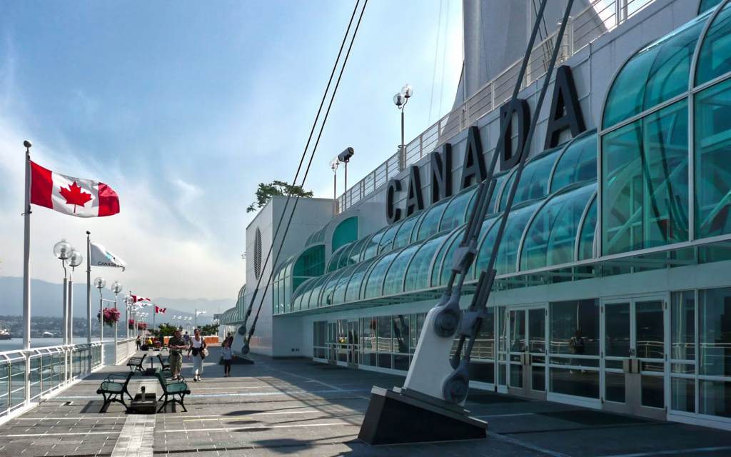 People walk along a waterfront promenade with a large "CANADA" sign, Canadian flags, benches, and glass-covered structures under a clear sky.