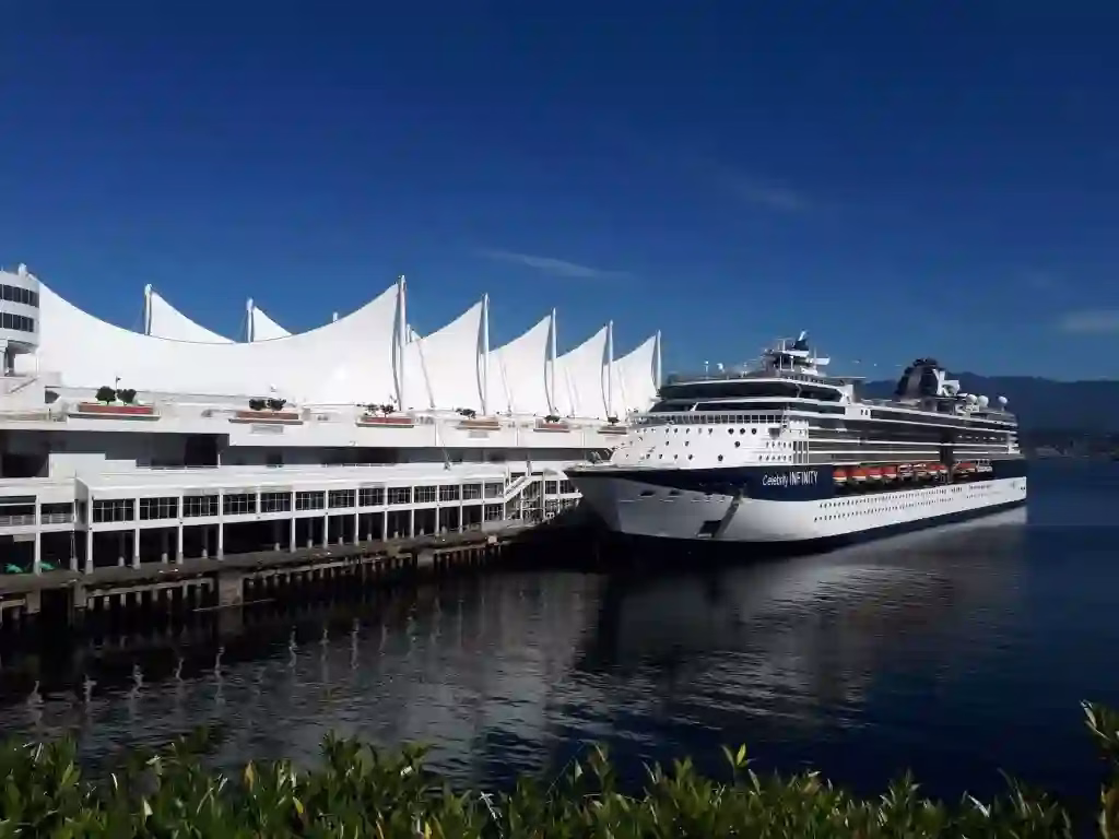 A large cruise ship docked at a modern white terminal with sail-like roof structures on a sunny day.