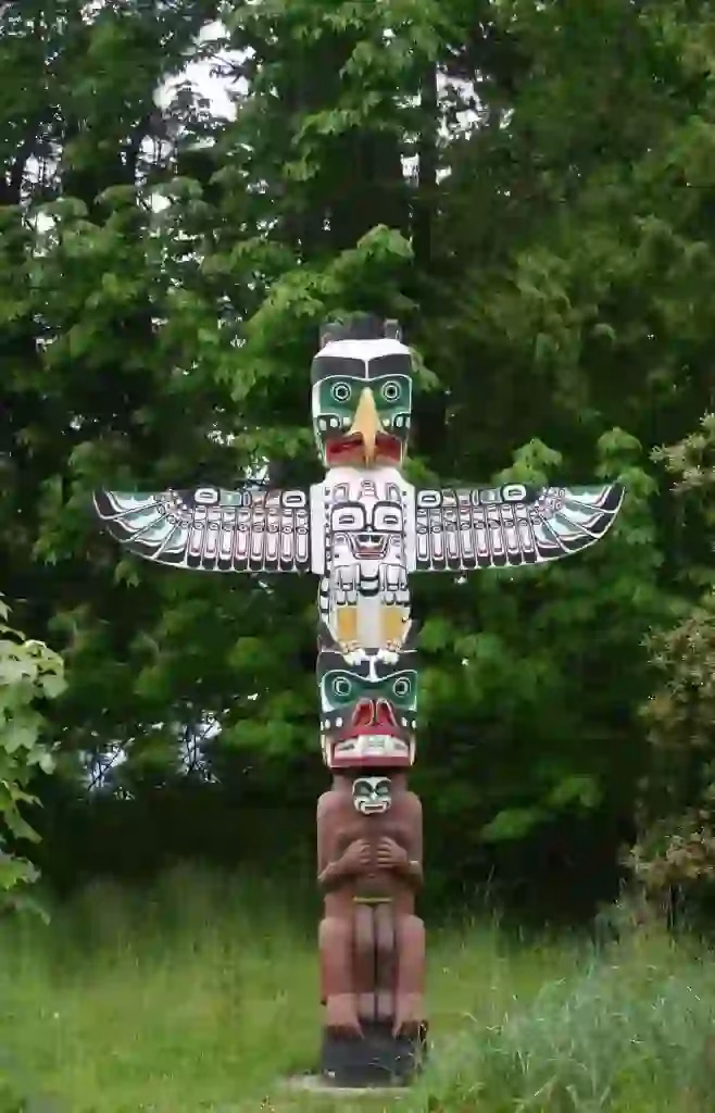 A painted totem pole with various animal and human figures stands in a grassy area surrounded by trees.